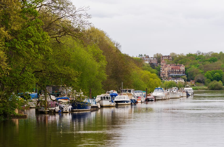 A panoramic view of a riverfront area during dusk, featuring calm water reflecting the evening sky and surrounding lights. Several small boats and yachts are moored along a concrete quay, with some bo